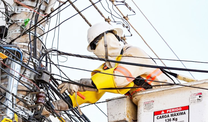 Poste urbano com técnicos desmontando fiação e removendo cabos com rua de centro ao fundo. - (Fotos: AGEMS)